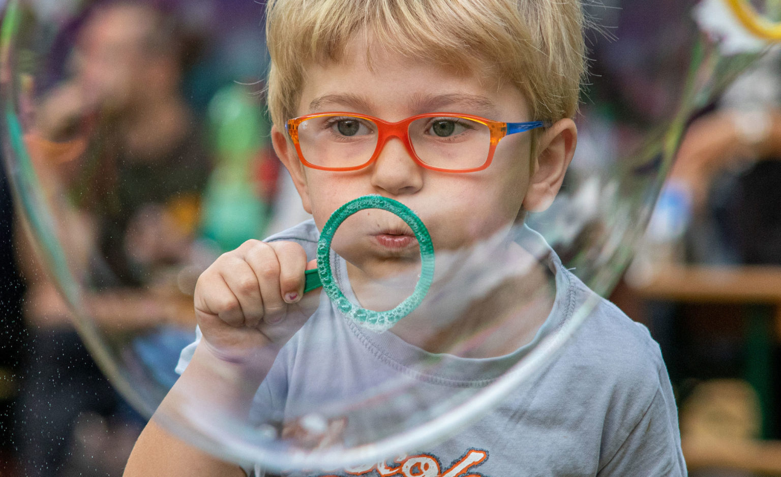 Ein kleiner Junge mit blondem Haar und orangefarbener Brille bläst Seifenblasen durch einen grünen Seifenblasenstab und konzentriert sich ganz konzentriert, während große Blasen vor seinem Gesicht schweben.