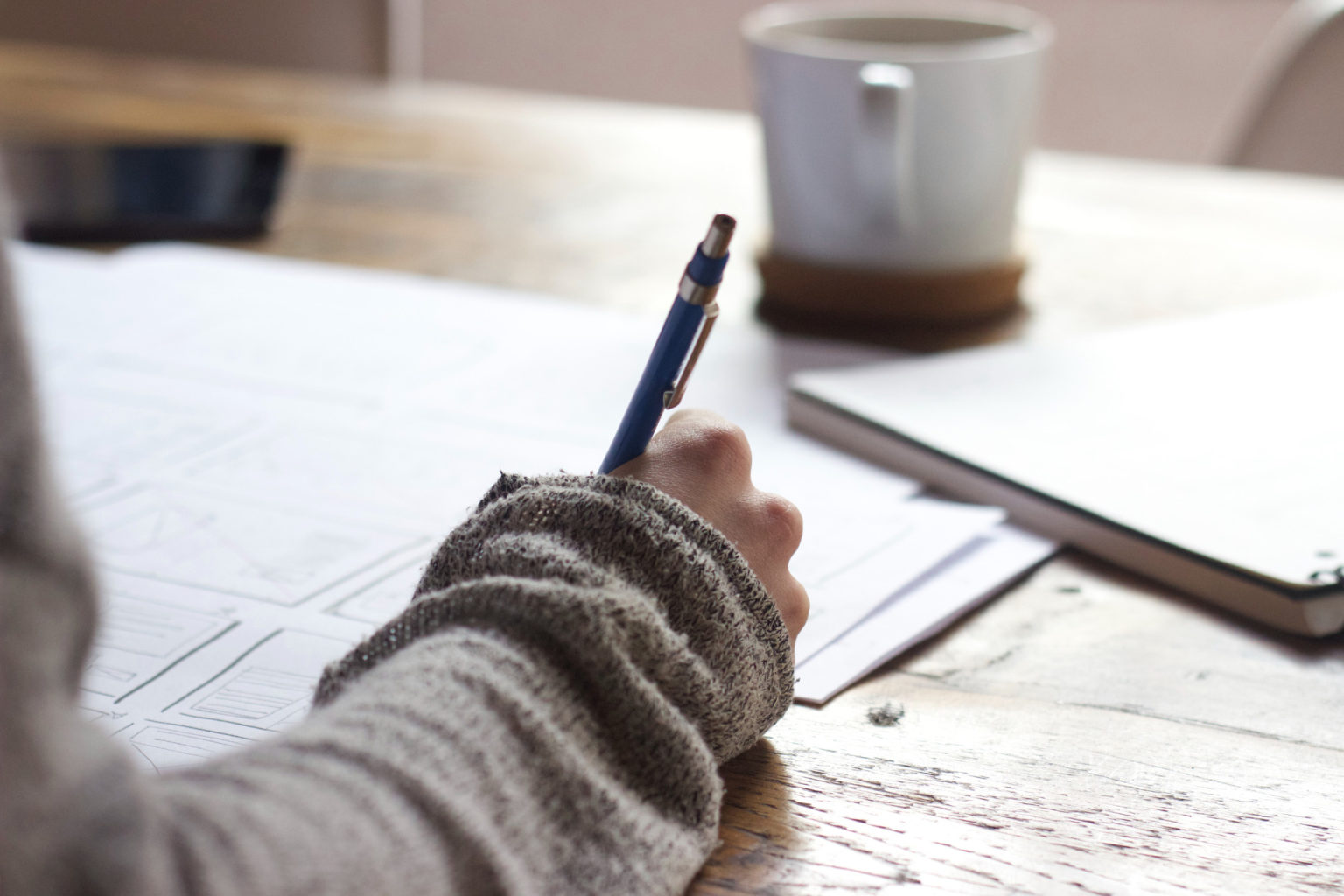 Eine Person in einem grauen Pullover schreibt mit einem Stift an einem Holztisch auf Papier. Auf dem Tisch stehen außerdem eine weiße Tasse und ein zugeklappter Laptop.