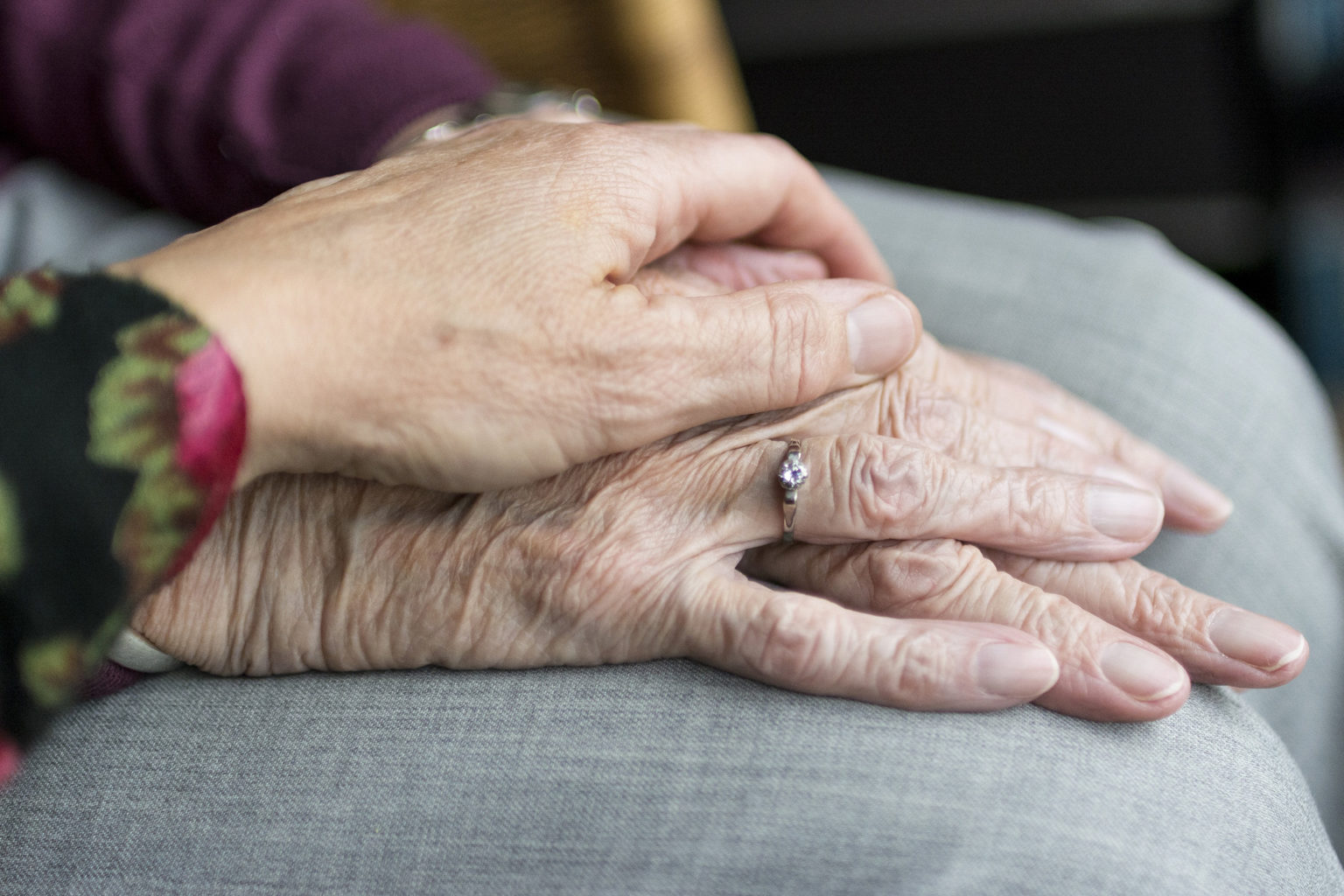 Eine jüngere Hand ruht sanft auf den Händen einer älteren Person und bietet Trost und Halt. Die ältere Person trägt einen silbernen Ring. Beide sitzen, ihre Hände ruhen auf einem grauen Stoff.