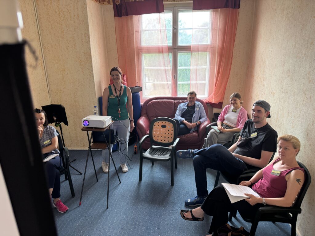 Sechs Personen sitzen in einem kleinen, gemütlichen Raum mit gelben Wänden und einem großen Fenster. Eine Person steht neben einem Projektor, die anderen sitzen auf Stühlen oder auf einem roten Sofa und blicken in die Kamera, als würden sie einen automatisch gespeicherten Entwurf betrachten.