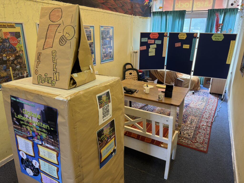 Ein gemütlicher, farbenfroher Raum mit Postern an gelben Wänden, einem Holztisch und einer Holzbank, blauen Schautafeln und einem großen Kasten mit der Aufschrift "automatisch gespeicherter Entwurf", der mit braunem Papier bedeckt ist, auf dem sich Schilder, ein QR-Code und Infoposter befinden. Sonnenlicht strömt durch tealfarbene Vorhänge.