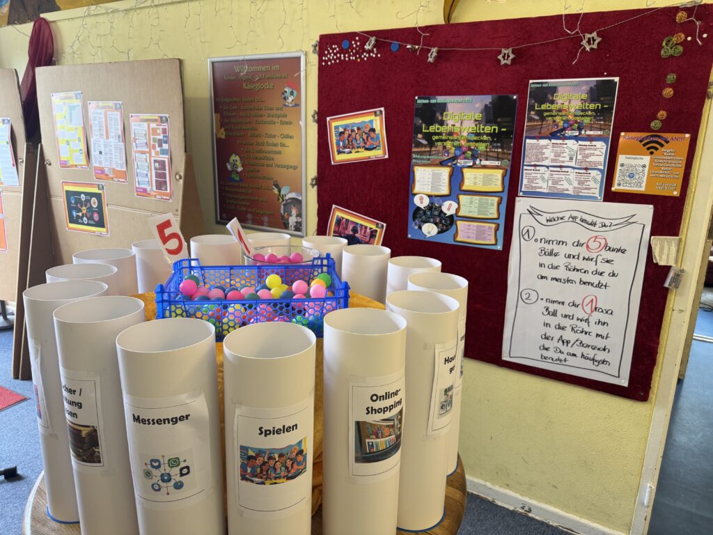 In einem Klassenzimmer sind auf einer roten Pinnwand Plakate zum Thema Internetsicherheit zu sehen. In der Mitte steht ein Kreis weißer zylindrischer Behälter, die mit Internetaktivitäten beschriftet sind, darunter automatisch gespeicherter Entwurf, und ein Korb mit bunten Plastikbällen.