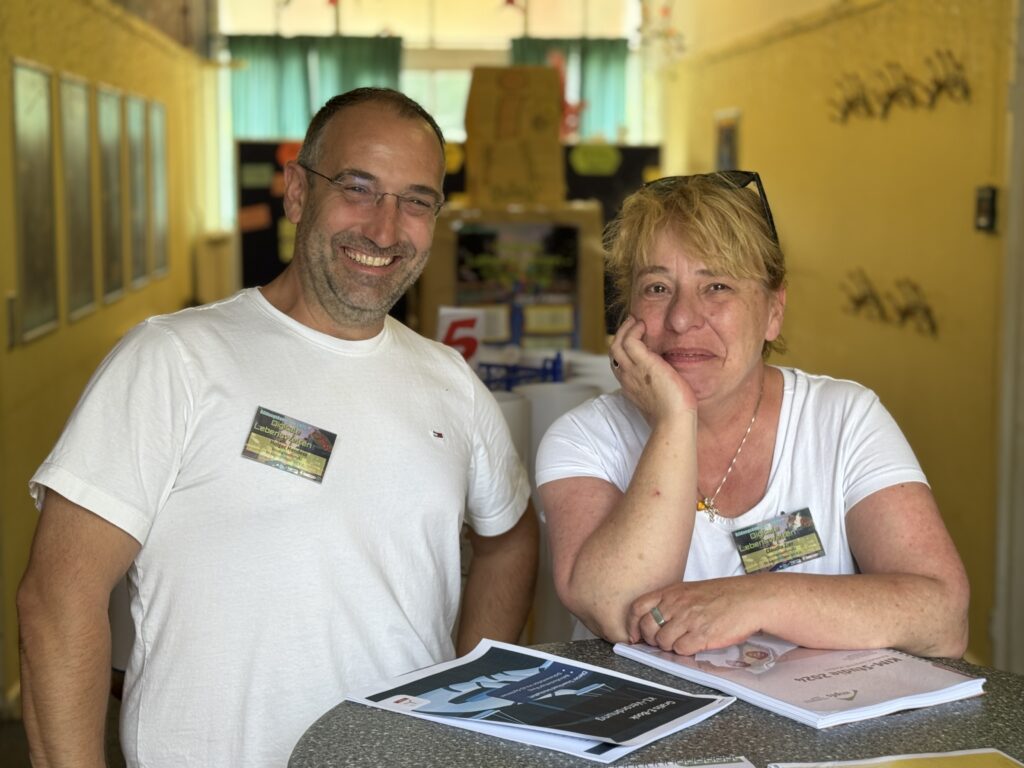 Zwei lächelnde Erwachsene, ein Mann und eine Frau, stehen an einem Tisch in einem hell erleuchteten Korridor. Sie tragen weiße Hemden und Namensschilder und sortieren Papiere mit der Aufschrift "automatisch gespeicherter Entwurf" vor sich. Die Atmosphäre ist freundlich und locker.