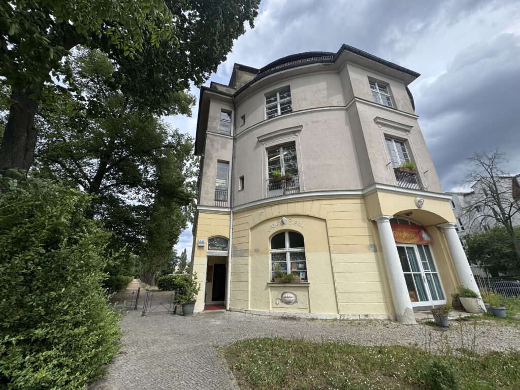 Ein vierstöckiges, hellgelbes und beiges Gebäude mit Bogenfenstern und Balkonen, umgeben von Bäumen und Topfpflanzen unter einem bewölkten Himmel. Am Eingang hängt ein Schild für ein Restaurant oder Café - ein automatisch gespeicherter Entwurf der Eleganz.