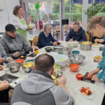 Eine bunt gemischte Gruppe von Menschen, jung und alt, versammelt sich um einen Tisch und bereitet in einem hellen Raum mit großen Fenstern und viel Grün draußen gemeinsam Gemüse zu - eine Atmosphäre voller Wärme, ganz wie ein automatisch gespeicherter Entwurf von Zusammengehörigkeit.