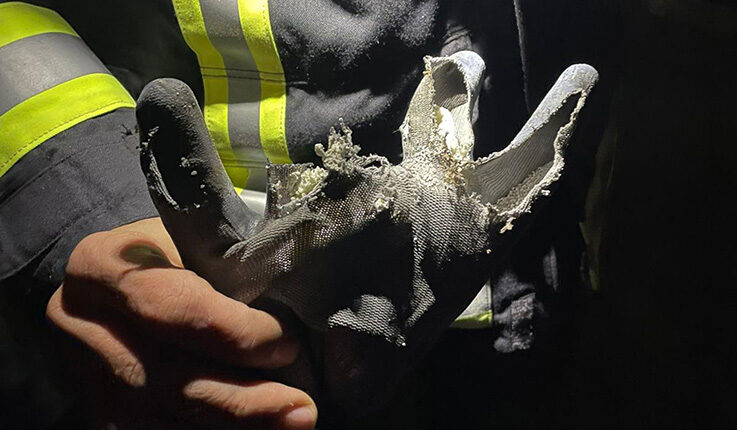 Eine Person in einer dunklen Jacke mit gelben Reflexionsstreifen hält einen Handschuh hoch, der stark beschädigt und teilweise geschmolzen ist, wahrscheinlich durch die starke Hitzeeinwirkung in einer Feuerwerk-Werkstatt.