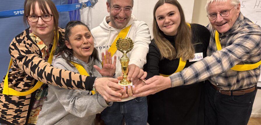 Fünf lächelnde Teammitglieder, die alle eine gelbe Schärpe tragen, stehen dicht beieinander und halten eine goldene Trophäe. Sie befinden sich in einem Raum und feiern ihre Leistung. Im Hintergrund sind ein Whiteboard und eine blaue Absperrung zu sehen.